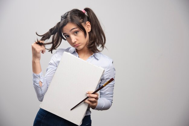 Young girl holding empty canvas and paintbrush on gray wall. 