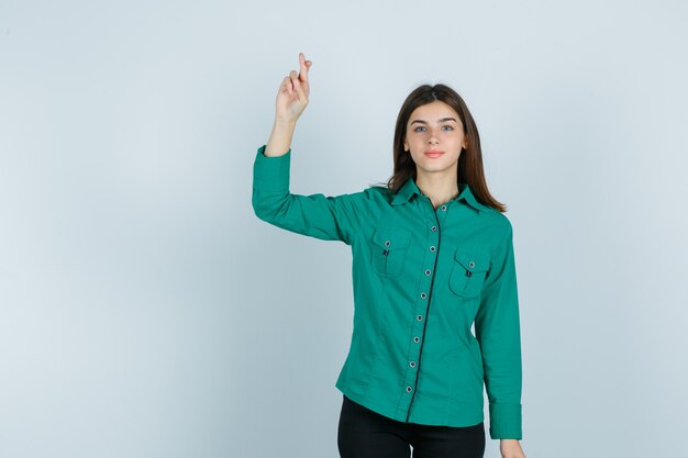 Young girl holding fingers crossed in green blouse, black pants and looking happy. front view.