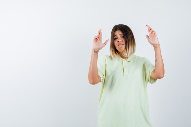 Young girl holding fingers crossed in t-shirt and looking worried. front view.