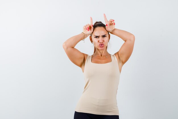 Young girl holding fingers over head as bull horns in beige top, black pants and looking amused , front view.