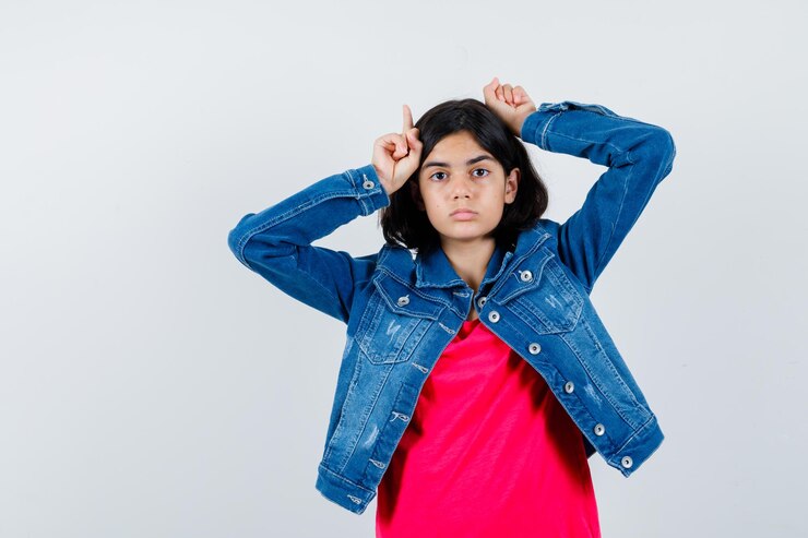 मुफ़्त फोटो लाल टी-शर्ट और जीन जैकेट में बैल के सींग के रूप में सिर के ऊपर उंगलियां रखती हुई प्यारी दिखती एक युवा लड़की।