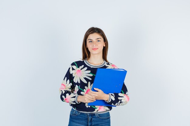 Young girl holding folder, looking at camera in floral blouse, jeans and looking cute , front view.