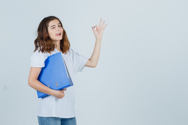 Young girl holding folder, showing ok gesture in white t-shirt and looking confident , front view.