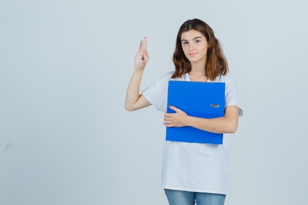 Young girl holding folder, showing ok gesture in white t-shirt and looking jolly , front view.