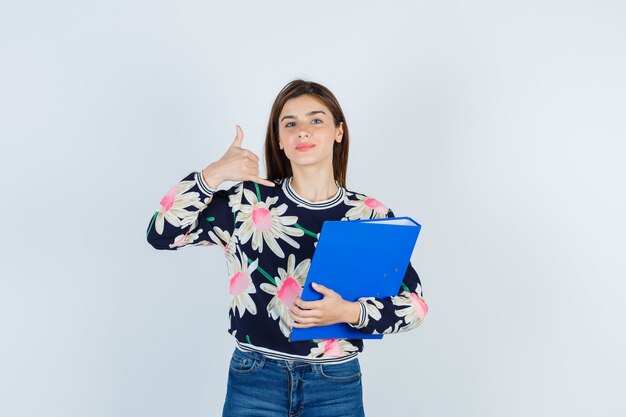 Young girl holding folder, showing phone gesture in floral blouse, jeans and looking confident , front view.