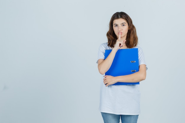 Молодая девушка держит папку, показывает жест молчания в белой футболке и выглядит уверенно. передний план.