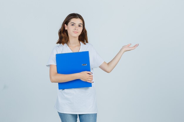 Young girl holding folder, showing welcoming gesture in white t-shirt and looking jolly , front view.