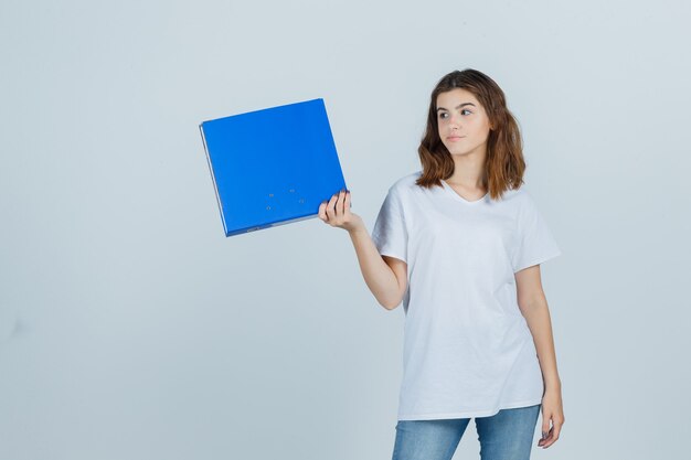 Young girl holding folder while looking aside in white t-shirt and looking displeased , front view.