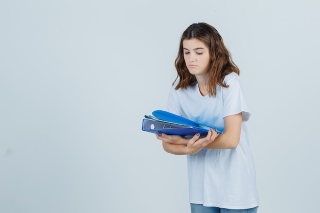 Молодая девушка держит папки в белой футболке и смотрит сосредоточенно, вид спереди.