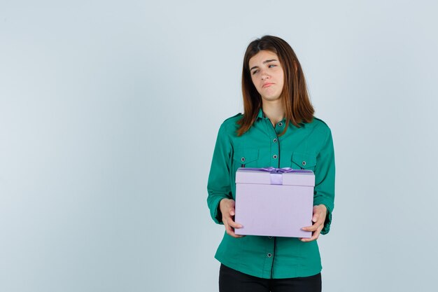 Young girl holding gift box in green blouse, black pants and looking morose. front view.