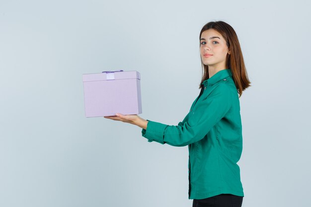 Young girl holding gift box, looking at camera in green blouse, black pants and looking happy , front view.