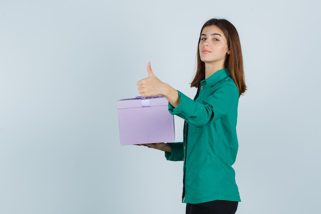 Молодая девушка держит подарочную коробку, показывает палец вверх в зеленой блузке, черных штанах и выглядит уверенно, вид спереди.