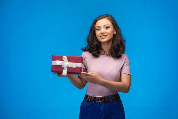 Молодая девушка держит подарочную коробку и улыбается на синем фоне.