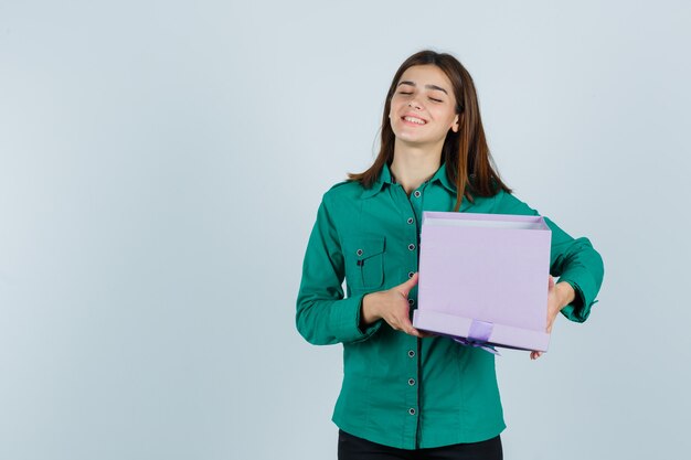 Young girl holding gift box, smiling in green blouse, black pants and looking merry , front view.