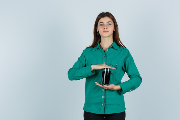 Young girl holding glass of black liquid in both hands in green blouse, black pants and looking cheery , front view.