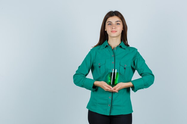 Молодая девушка держит стакан зеленой жидкости обеими руками в зеленой блузке, черных штанах и выглядит весело, вид спереди.