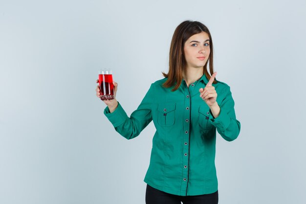 Young girl holding glass of red liquid, showing warning gesture in green blouse, black pants and looking cute , front view.