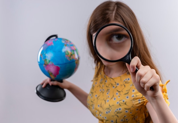 Foto gratuita ragazza che tiene il globo e guardando attraverso la lente di ingrandimento sul muro bianco isolato