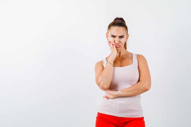 Young girl holding hand on cheek, grimacing in beige top and red pants and looking serious , front view.