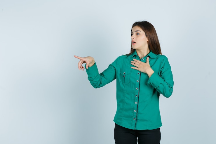 मुफ़्त फोटो young girl holding hand over chest, pointing left with index finger in green blouse, black pants and looking surprised , front view.