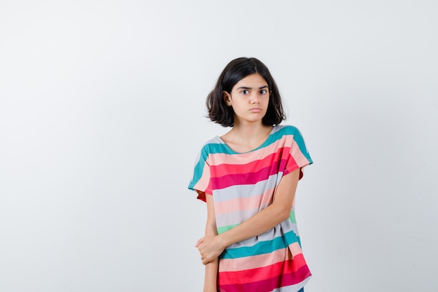Молодая девушка держит руку на предплечье в красочной полосатой футболке и выглядит серьезным, вид спереди.