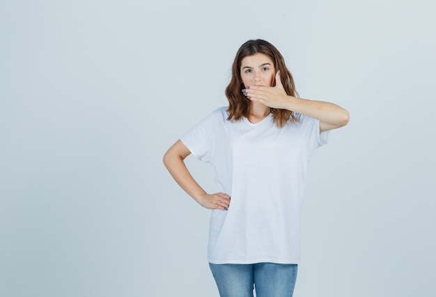 Young girl holding hand on mouth in white t-shirt and looking surprised , front view.