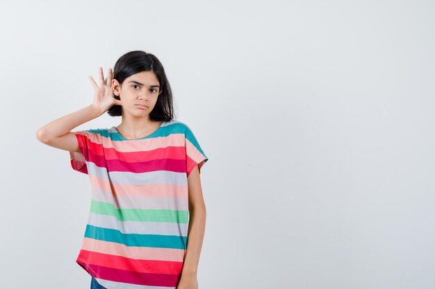 Young girl holding hand near ear to hear in colorful striped t-shirt and looking focused. front view.