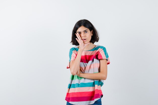 Young girl holding hand near mouth as telling secret in colorful striped t-shirt and looking surprised. front view.
