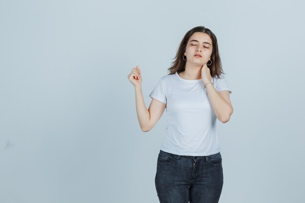 Young girl holding hand on neck in t-shirt, jeans and looking unwell , front view.