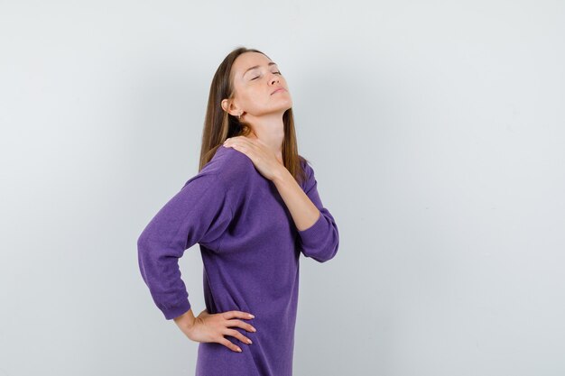 Молодая девушка держит руку на плече в фиолетовой рубашке и выглядит расслабленной, вид спереди.