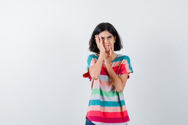 Молодая девушка держит руки на щеке и подбородке в красочной полосатой футболке и выглядит задумчиво. передний план.