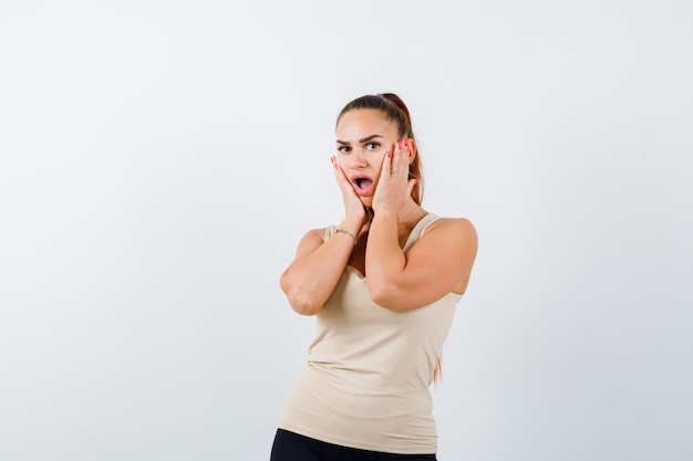 Young girl holding hands on cheeks in beige top, black pants and looking surprised