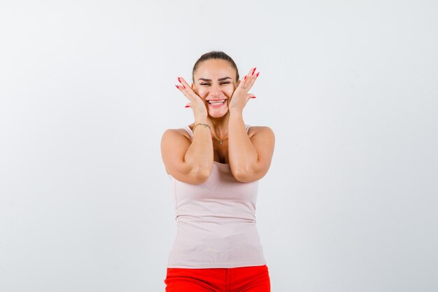 Young girl holding hands on cheeks in beige top and red pants and looking happy , front view.