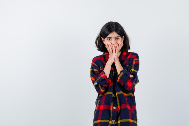 Young girl holding hands on cheeks in checked shirt and looking timid. front view.