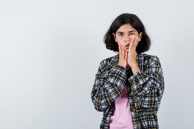 Young girl holding hands on cheeks in checked shirt and pink t-shirt and looking surprised. front view.