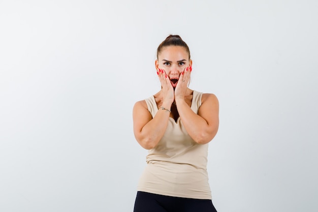 Young girl holding hands on cheeks, keeping mouth wide open in beige top, black pants