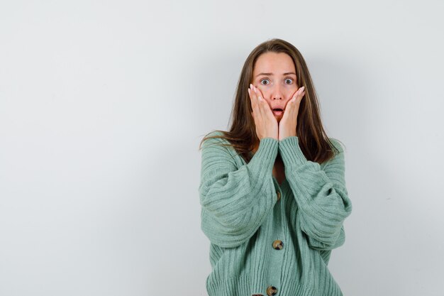 Young girl holding hands on cheeks in knitwear and looking surprised , front view.