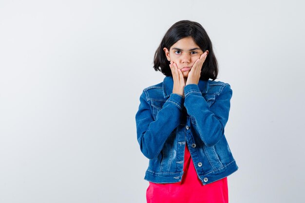 Young girl holding hands on cheeks in red t-shirt and jean jacket and looking serious. front view.