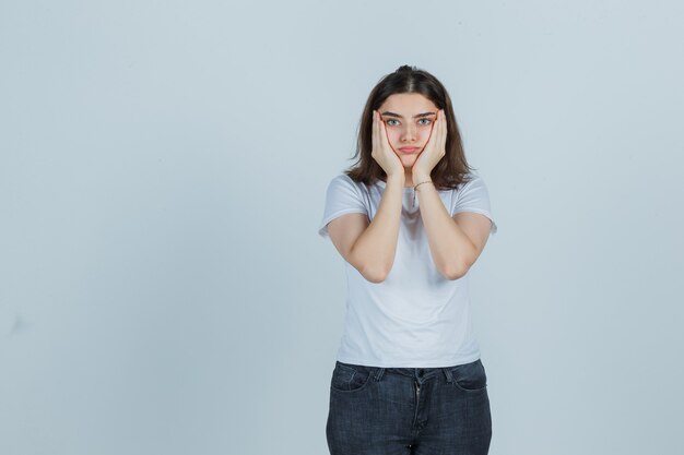 Young girl holding hands on cheeks in t-shirt, jeans and looking puzzled , front view.