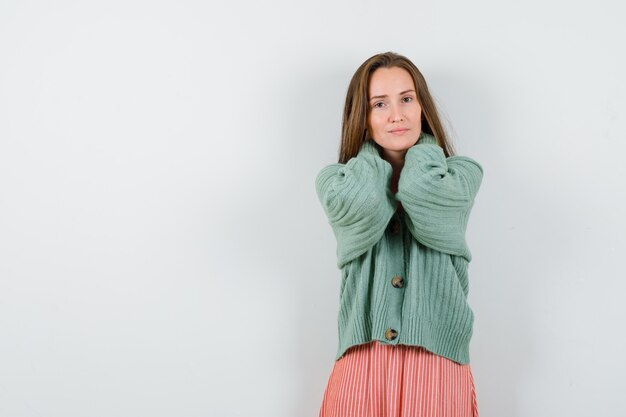 Young girl holding hands behind head in knitwear, skirt and looking serious. front view.