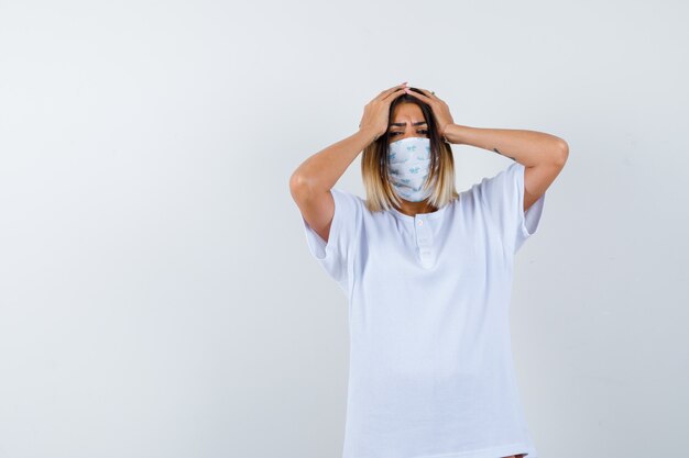 Young girl holding hands on head in white t-shirt and mask and looking worried. front view.