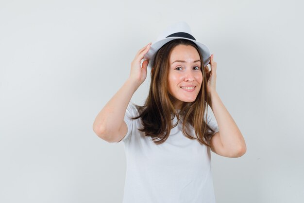Молодая девушка держит руки на шляпе в белой футболке и выглядит довольно. передний план.