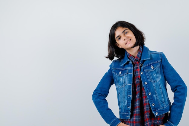 Young girl holding hands on waist, bending left in checked shirt and jean jacket and looking happy. front view.