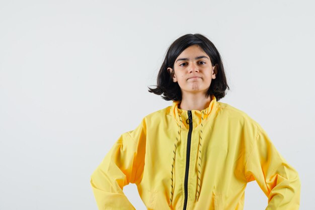 Young girl holding hands on waist in yellow bomber jacket and looking serious.