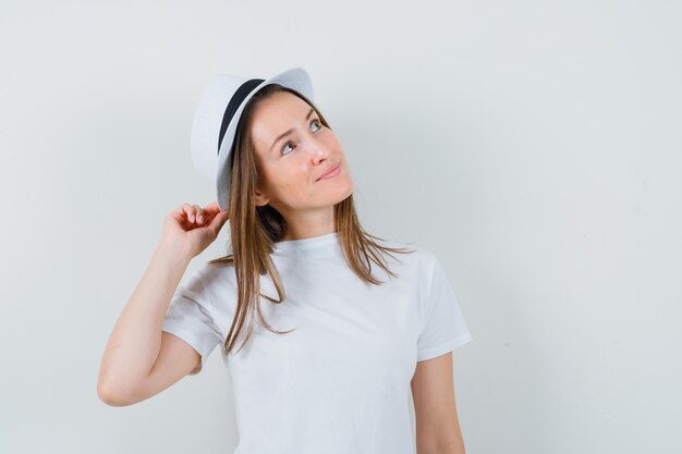 Young girl holding hat while looking up in white t-shirt and looking dreamy. front view.