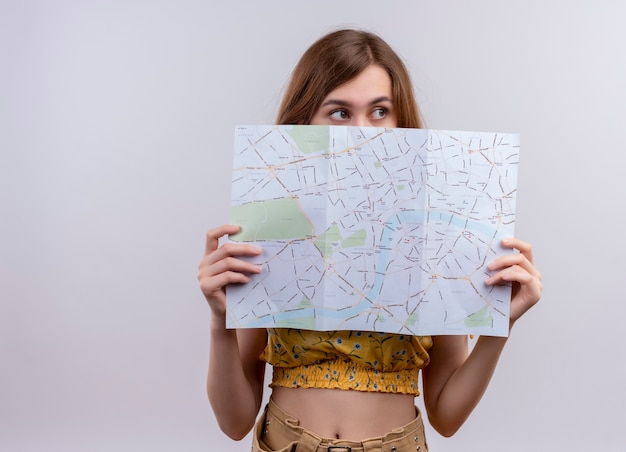 Young girl holding map hiding behind it and looking at right side on isolated white wall with copy space