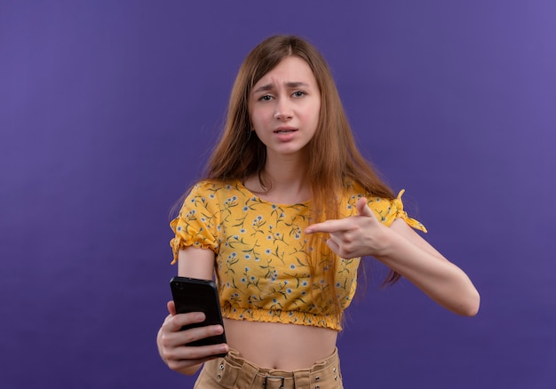 Free photo young girl holding mobile phone and pointing at it on isolated purple wall with copy space