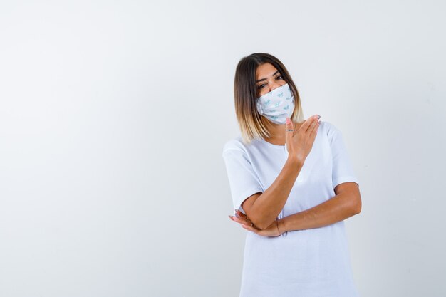 Young girl holding one hand under elbow, stretching hand as holding something in white t-shirt and mask and looking serious , front view.