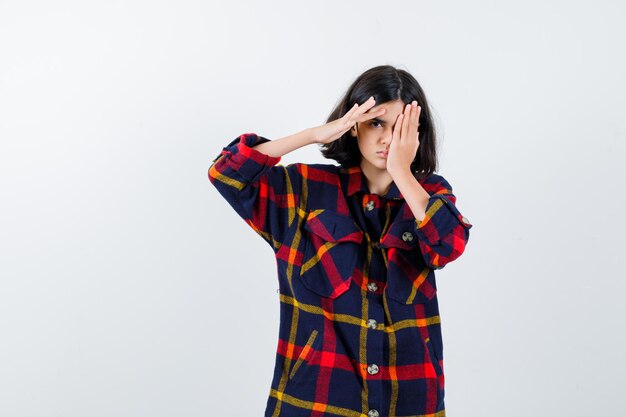 Young girl holding one hand on forehead, another hand on eye in checked shirt and looking serious , front view.