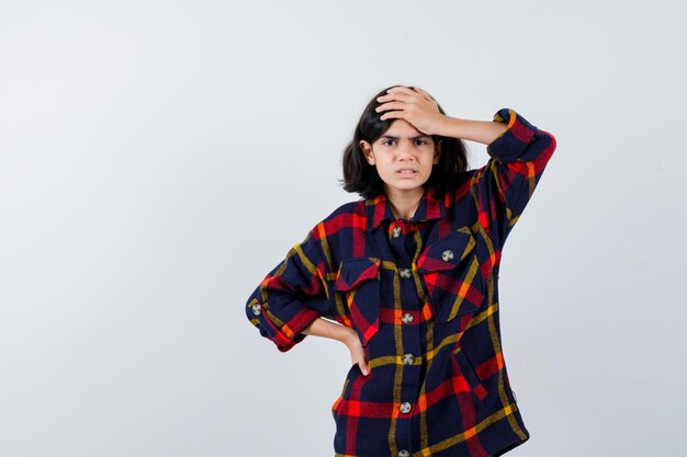 Young girl holding one hand on forehead while holding another hand on waist in checked shirt and looking annoyed , front view.